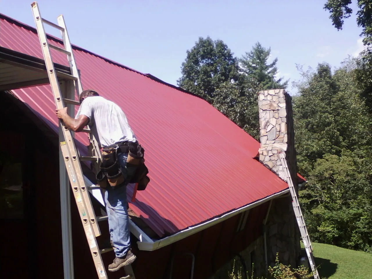Roofer on a ladder beside a red metal roof and stone chimney — Soffit & Fascia Repair by Roofing Brothers in Boxford